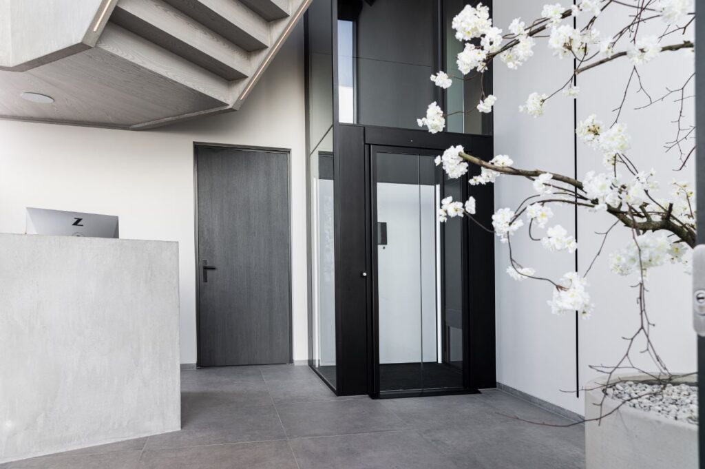 Zwarte glazen lift in een luxe entree met betonnen balie en bloesemboom.