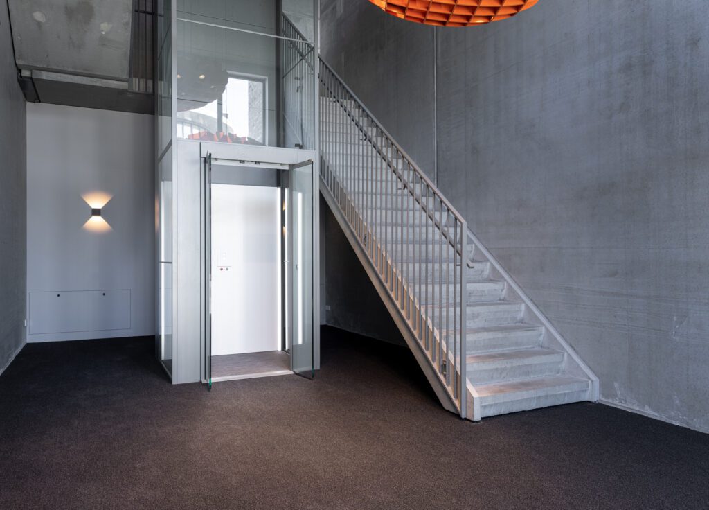Moderne glazen lift met geopende deur naast een betonnen trap in een ruim trappenhuis met grijze wanden.