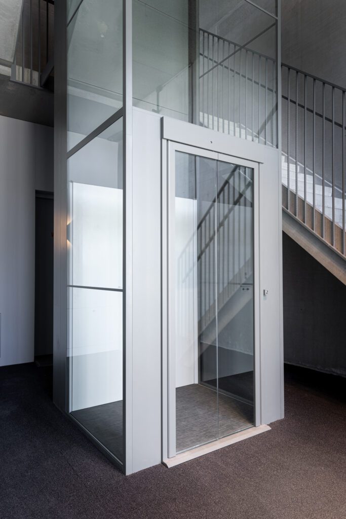 Detail van een glazen lift met aluminium frame naast een betonnen trap.