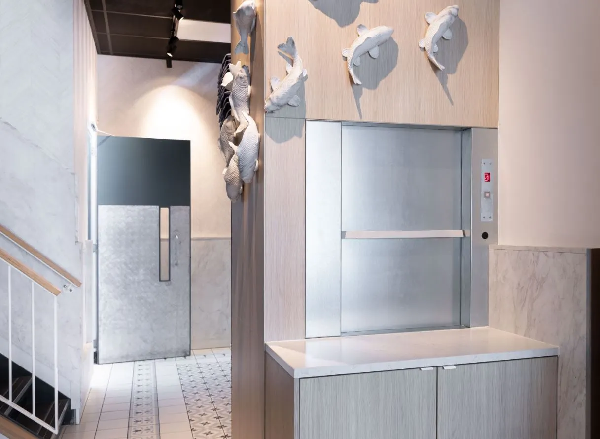 Kleine goederenlift met roestvrijstalen schuifdeur in een licht interieur, omringd door houten panelen en decoratieve vissen aan de muur.