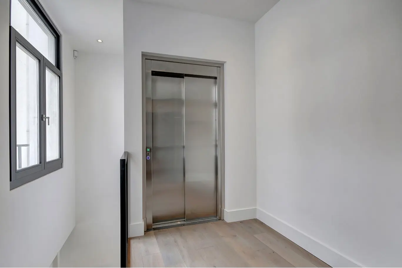 Heldere, moderne gang met houten vloer en een roestvrijstalen liftdeur naast een groot raam met zwarte omlijsting.