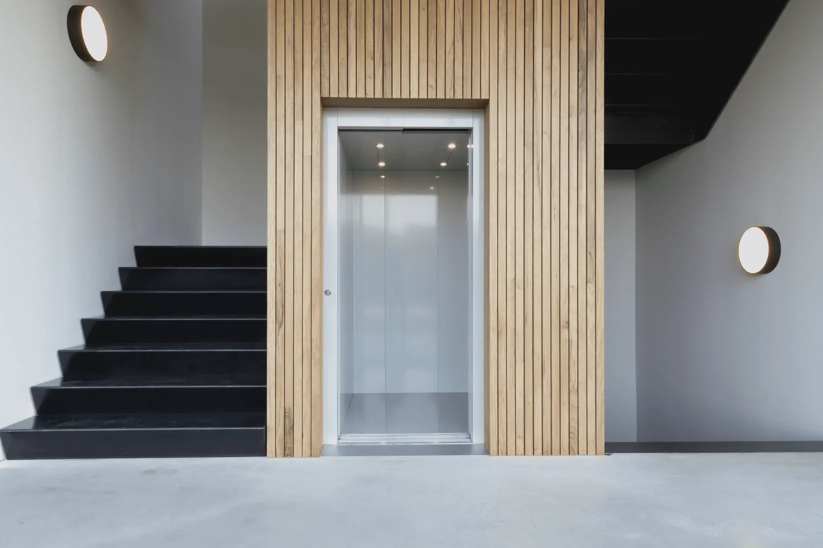 Moderne lift met witte deuren ingebouwd in een wand van verticale houten latten, naast een brede zwarte trap in een minimalistische ruimte.