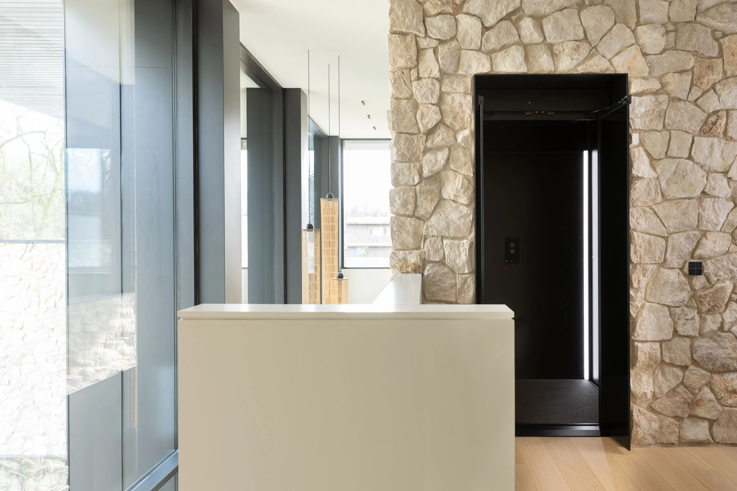 Moderne zwarte lift ingebouwd in een natuurstenen muur, binnen een licht en ruim interieur met grote ramen en houten vloer.